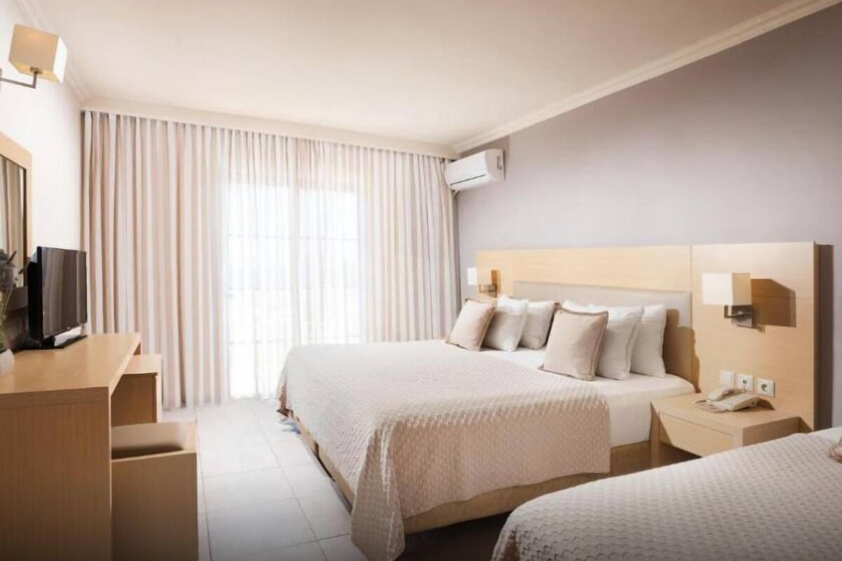Habitación de hotel con dos camas, un escritorio con televisor, una mesita de noche con teléfono y un ventanal con cortinas.