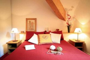 Una cama bien tendida con ropa de cama roja, dos lámparas de noche, un florero con flores y un cuadro en la pared.