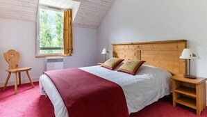 Een slaapkamer met een houten hoofdeinde, een bed met witte en rode beddengoed, een nachtkastje met een lamp, een stoel en een raam met gordijnen.