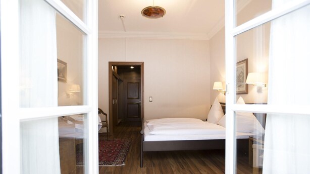 Quarto com cama, piso de madeira, porta que dá para outro cômodo e um tapete pequeno.