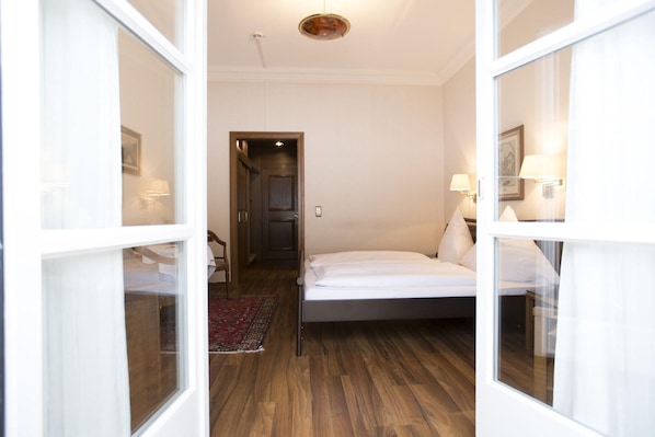 A bedroom with a bed, a wooden floor, a door leading to another room, and a small rug. - Mueller (Schwangau)
