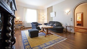 Uma sala de estar com sofá, pufe e mesa de centro, uma escrivaninha com cadeira, um radiador e uma janela com cortinas.