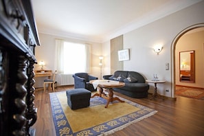 A living room with a sofa, ottoman, and coffee table, a desk with a chair, a radiator, and a window with curtains. - Mueller (Schwangau)