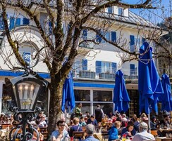 Un café al aire libre con clientes sentados en mesas, un árbol grande en primer plano y un edificio con contraventanas azules al fondo.