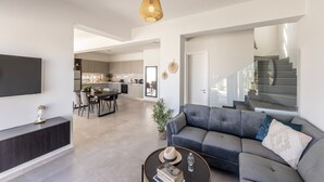 Un soggiorno moderno con un divano grigio, un tavolino e una televisione a schermo piatto. Si nota una scala che conduce a un piano superiore, e sullo sfondo si intravede un angolo cottura.
