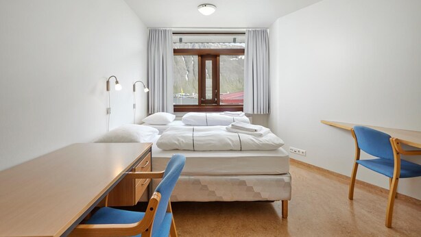 A small, clean room with a bed, a desk, and a chair. There is a window with curtains, a lamp, and a view of a building outside. - Edda Hotel Isafjorour (Isafjordur)