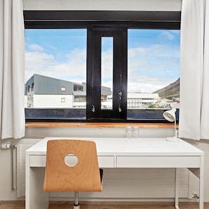 Una habitación con un escritorio blanco, una silla de madera, una ventana con vistas a edificios y una lámpara blanca.