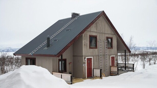 Una casa a due piani in legno, con tetto spiovente, un portico e una porta rossa.