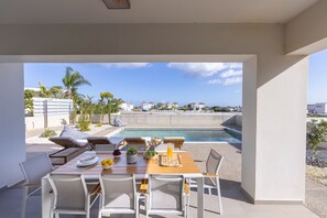 Un espace repas extérieur avec vue sur la piscine, doté de mobilier blanc et bénéficiant d’un ciel dégagé.