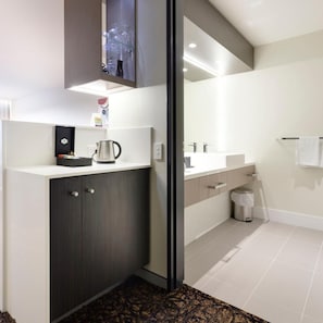 A modern bathroom with a dark wood cabinet, a glass shelf with toiletries, a coffee maker, and a large mirror. - Calamvale Hotel Suites And Conference Centre (Brisbane)