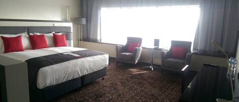 A hotel room with a large bed, two armchairs, a desk, and a large window with curtains.