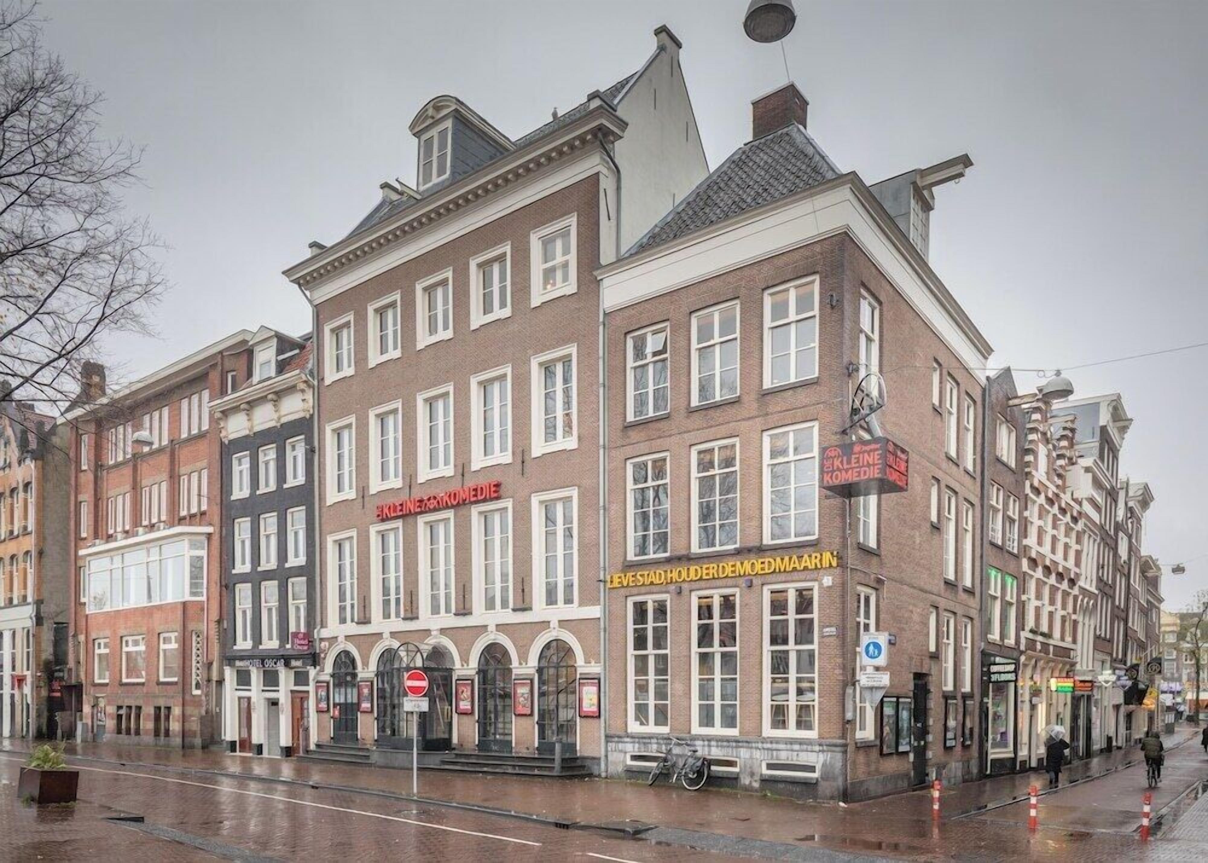 A historic building with multiple stories, featuring a prominent red sign for 'Kleine Komedie' and another sign for 'Leve Stad, Honder Demoeed Maand In'.