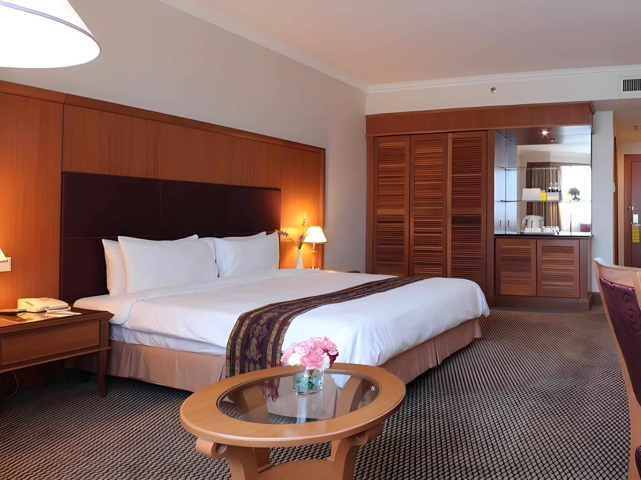 A hotel room with a large bed, wooden headboard, a round glass table with a vase of flowers, a nightstand with a telephone, and a wardrobe with sliding doors.