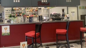 Een bar met een rode toonbank, twee rode barkrukken en diverse flessen en glazen op planken.