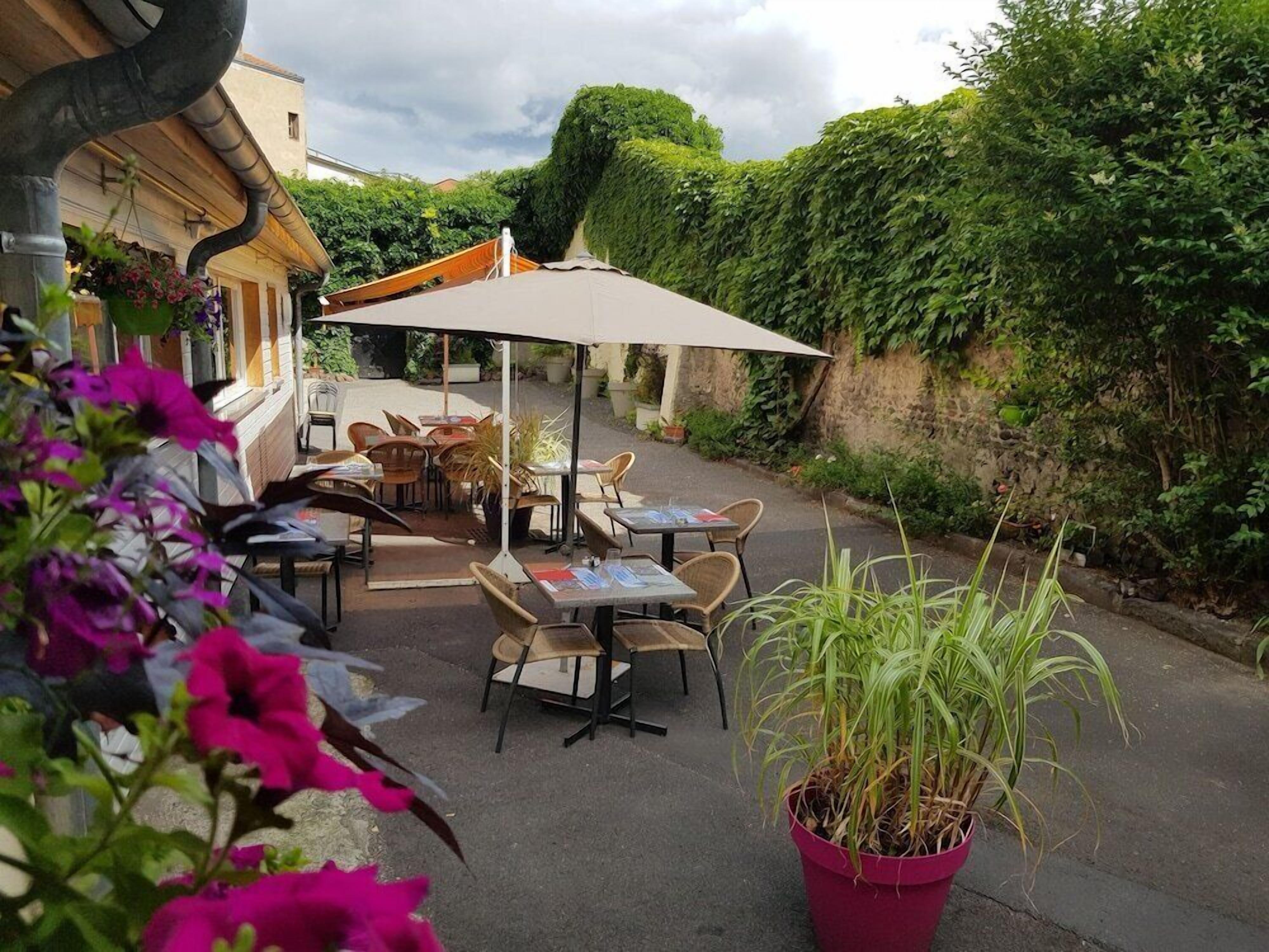 An outdoor cafe with tables and chairs, surrounded by greenery and a stone wall.
