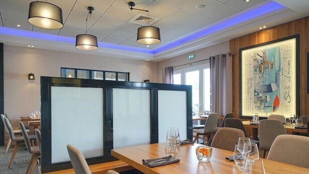 A modern conference room with a large table, chairs, and a glass partition. There are ceiling lights, a framed artwork on the wall, and a window with curtains. - Brit Hotel Restaurant Le Cottage (Arnage)