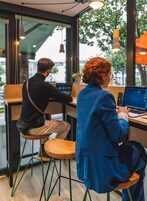Deux personnes travaillent à une table en bois, devant un ordinateur portable, dans un café moderne doté de grandes fenêtres et d’un éclairage suspendu.