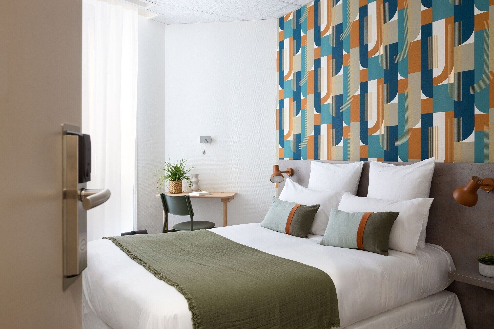 A hotel room with a bed, a desk, and a chair. There is a wall with a geometric pattern.
