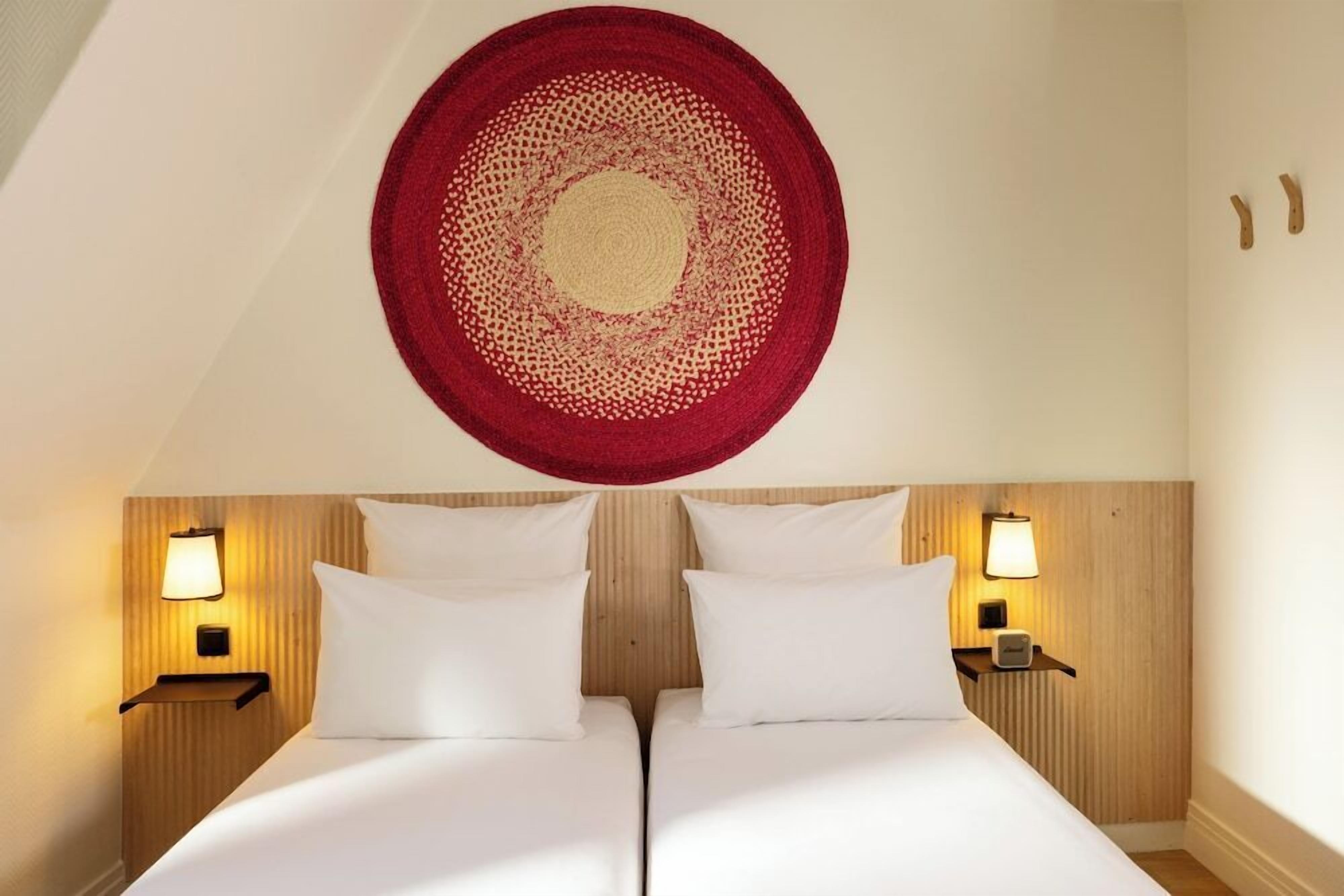 A hotel room with two beds, wooden headboard, wall-mounted lamps, and a circular wall hanging.