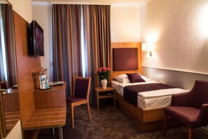 Ein Hotelzimmer mit einem Bett, einem Schreibtisch mit Stuhl, einem Fernseher, einem Nachttisch mit Blumenvase und einem großen Fenster mit Vorhängen.