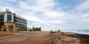 Eine Küstenstraße mit einem modernen Gebäude links, einem Sandstrand mit verstreuten Felsen und dem Meer im Hintergrund.