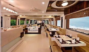 A restaurant with a view of the ocean, featuring a long counter, multiple dining tables, and a sign that reads "Blue Coriander the Restaurant". - Lords Eco Inn Porbandar (Porbandar)