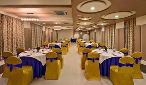 A banquet hall with round tables, yellow chairs, and blue sashes. - Lords Eco Inn Porbandar (Porbandar)