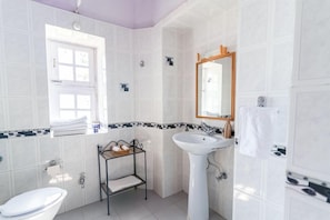 A bathroom with white tiles, a pedestal sink, a vanity with a mirror, and a window with a view. - Tree of Life Grand Oak Manor (Almora)