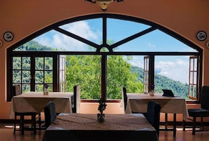 A dining area with a table set for two, large windows with a view of a forested mountain range, and a ceiling with a decorative element. - Tree of Life Grand Oak Manor (Almora)