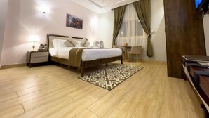 A hotel room with a bed, bedside tables, a desk, a chair, a TV, and a patterned rug. - Landmark Hotel Suites (Tabuk)