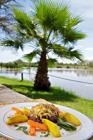 A plate of food with vegetables and meat, with a palm tree and water body in the background. - Voyager Ziwani Tented Camp (Kenya)