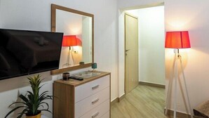 Habitación de hotel con televisión, escritorio con cajones, refrigerador pequeño, planta en maceta, espejo y lámpara.