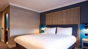 Quarto de hotel com uma cama grande, cabeceira de madeira, duas luminárias fixadas na parede e uma pequena mesa de cabeceira.