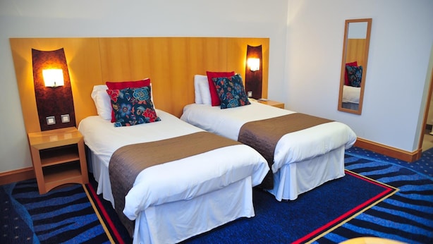 Een hotelkamer met twee bedden, een houten hoofdbord, nachtlampjes, een spiegel en een blauw tapijt.
