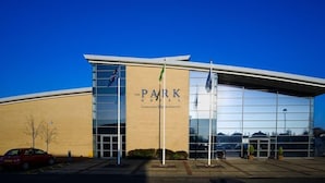 Een modern gebouw met een grote glazen gevel, met de naam 'The Park Hotel', en een opvallende ingang en een vlaggenmast.