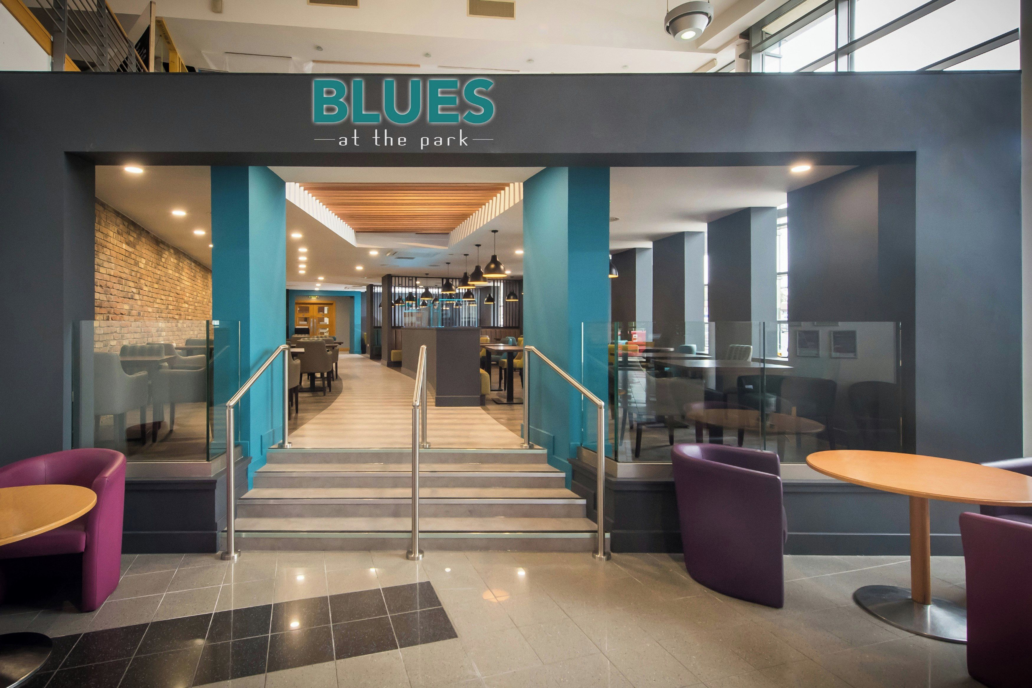 A modern interior with a staircase leading to a seating area, a sign reading 'BLUES at the park', and a mix of seating options including tables and chairs.