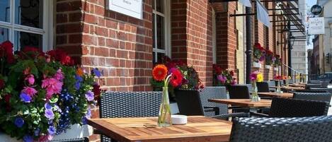 Outdoor seating with tables and chairs, a brick building, and flower arrangements.