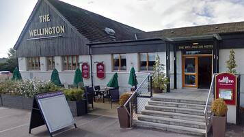 A hotel with a sign reading 'The Wellington Hotel' and an adjacent 'Eating Inn' sign. There are outdoor seating areas with umbrellas and a staircase leading to the entrance.