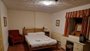 Ein Schlafzimmer mit einem hölzernen Bett, einem roten Sessel, einem Nachttisch mit Lampe, einer Kommode und einem Spiegel.