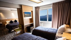 Une chambre d’hôtel avec deux lits, une tête de lit en bois, un petit bureau, une chaise, une fenêtre donnant sur les montagnes et un tableau accroché au mur.