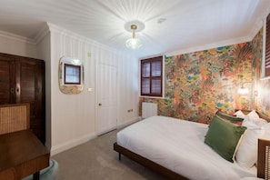 A bedroom with a bed, a dresser, a mirror, and a wall with floral wallpaper. - The Clarence (Portsmouth)