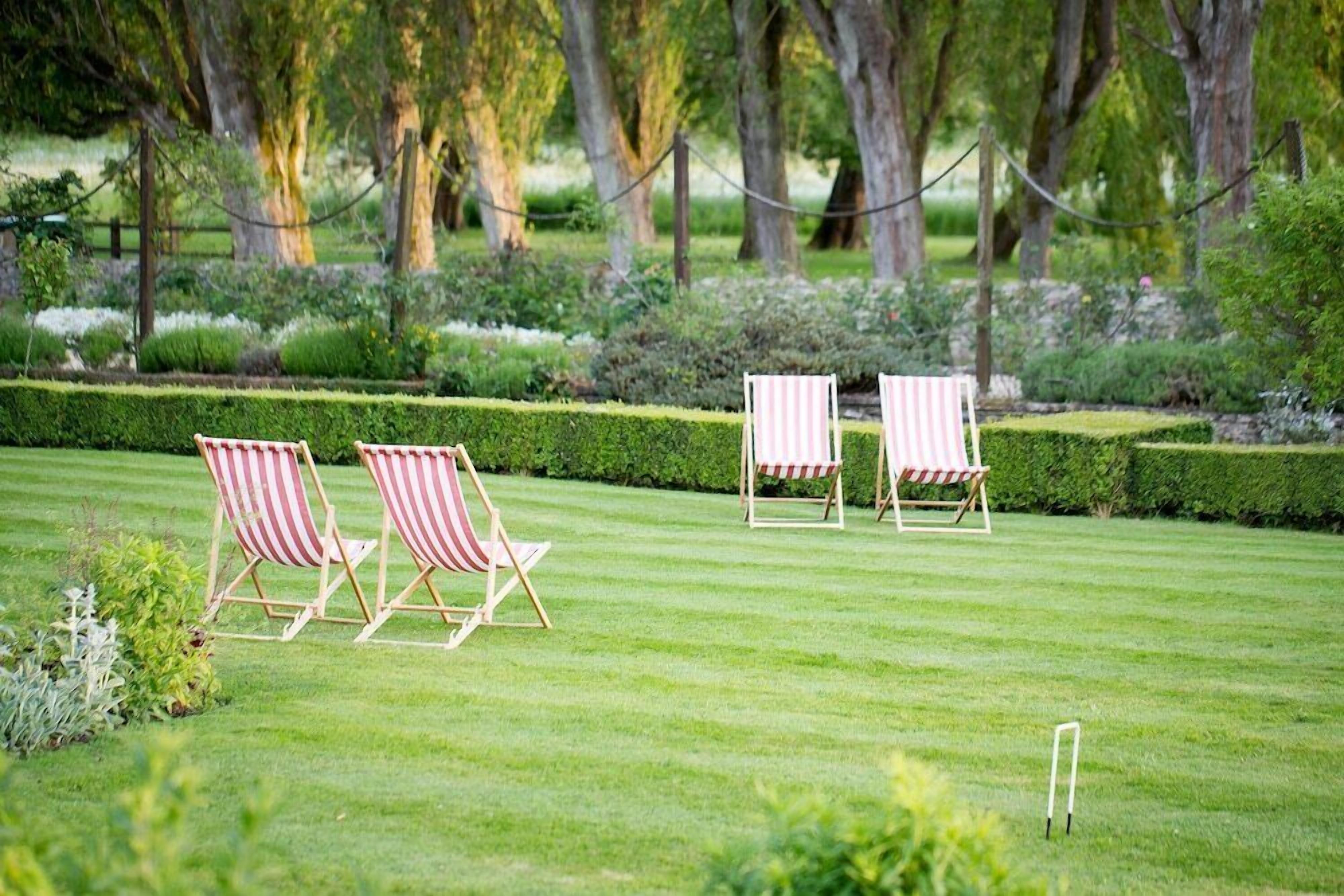 A well-manicured lawn with three striped lawn chairs and a small table, surrounded by neatly trimmed hedges and trees.