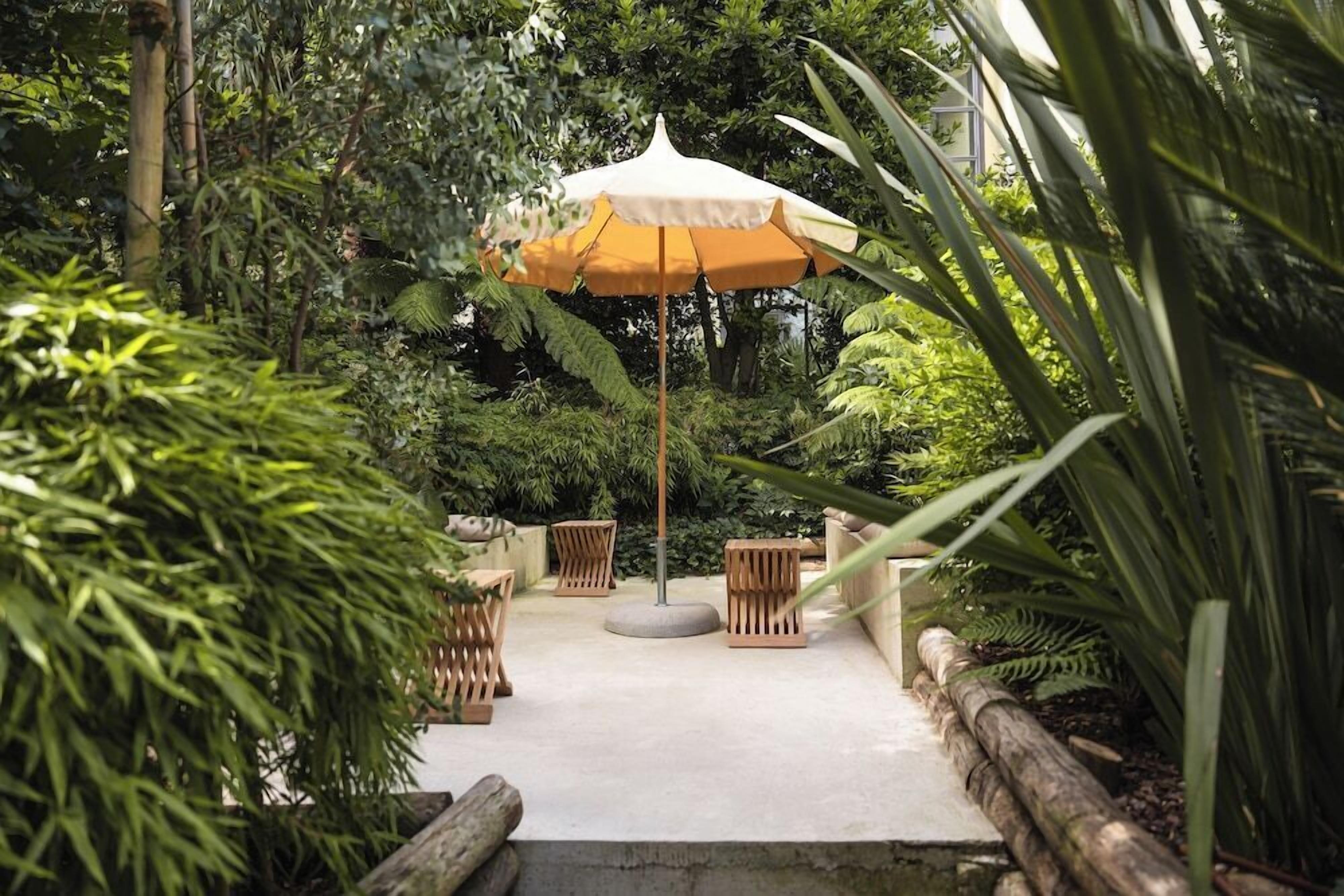 A garden patio with a central umbrella, surrounded by wooden furniture and lush greenery.