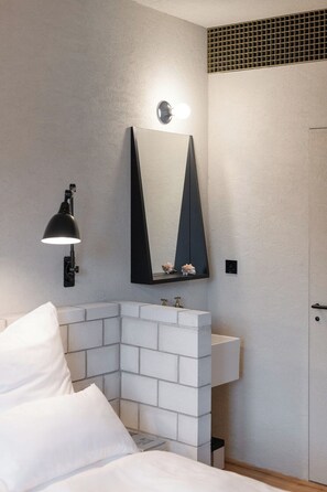 Une salle de bain moderne avec un mur en briques blanches, un miroir à cadre noir, un lavabo blanc et une lampe noire fixée au mur.