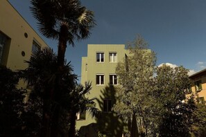 Un edificio moderno con múltiples ventanas y techo plano, rodeado de árboles y otros edificios.