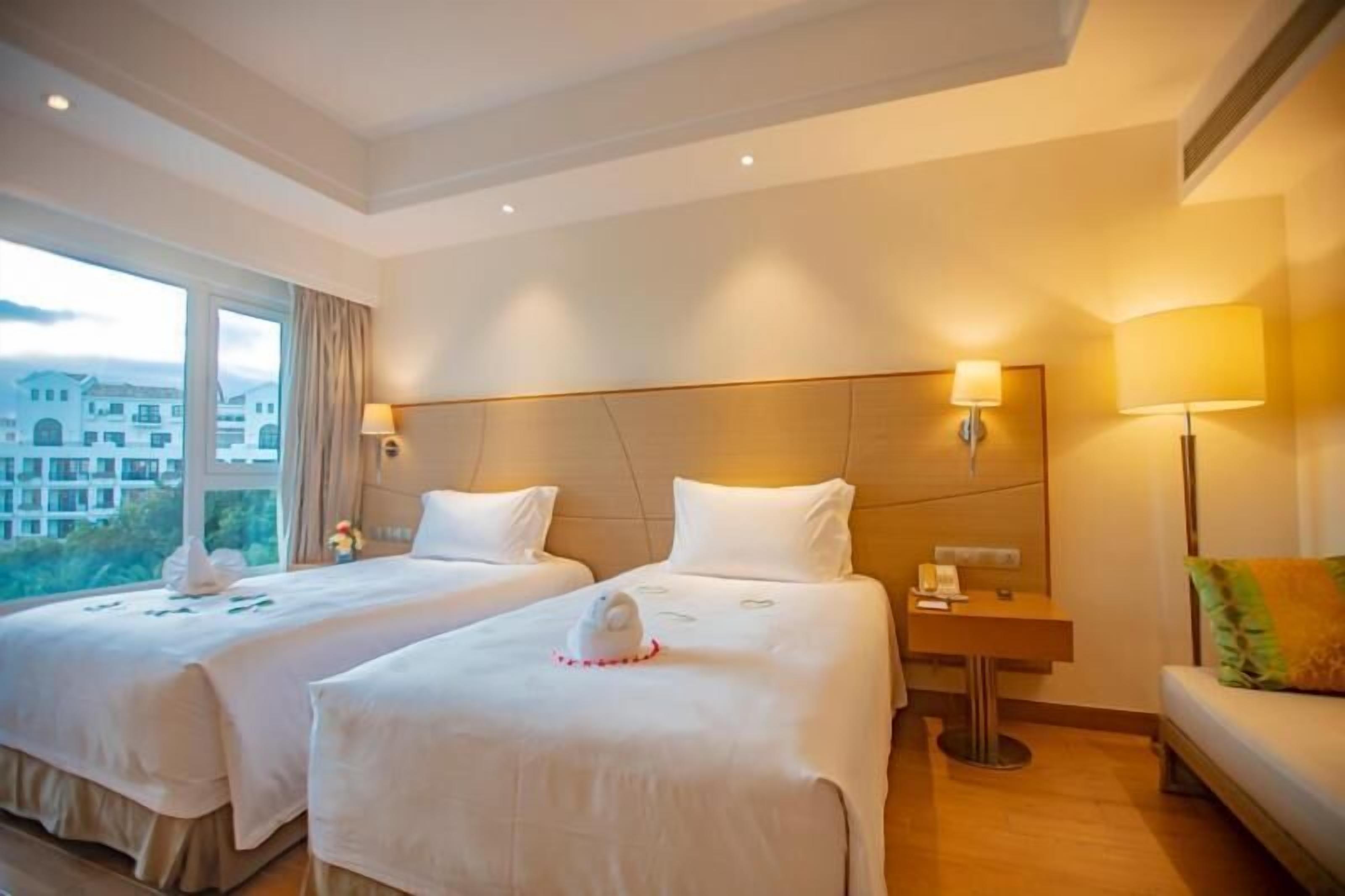 A hotel room with two beds, a nightstand, a lamp, and a view of buildings outside.