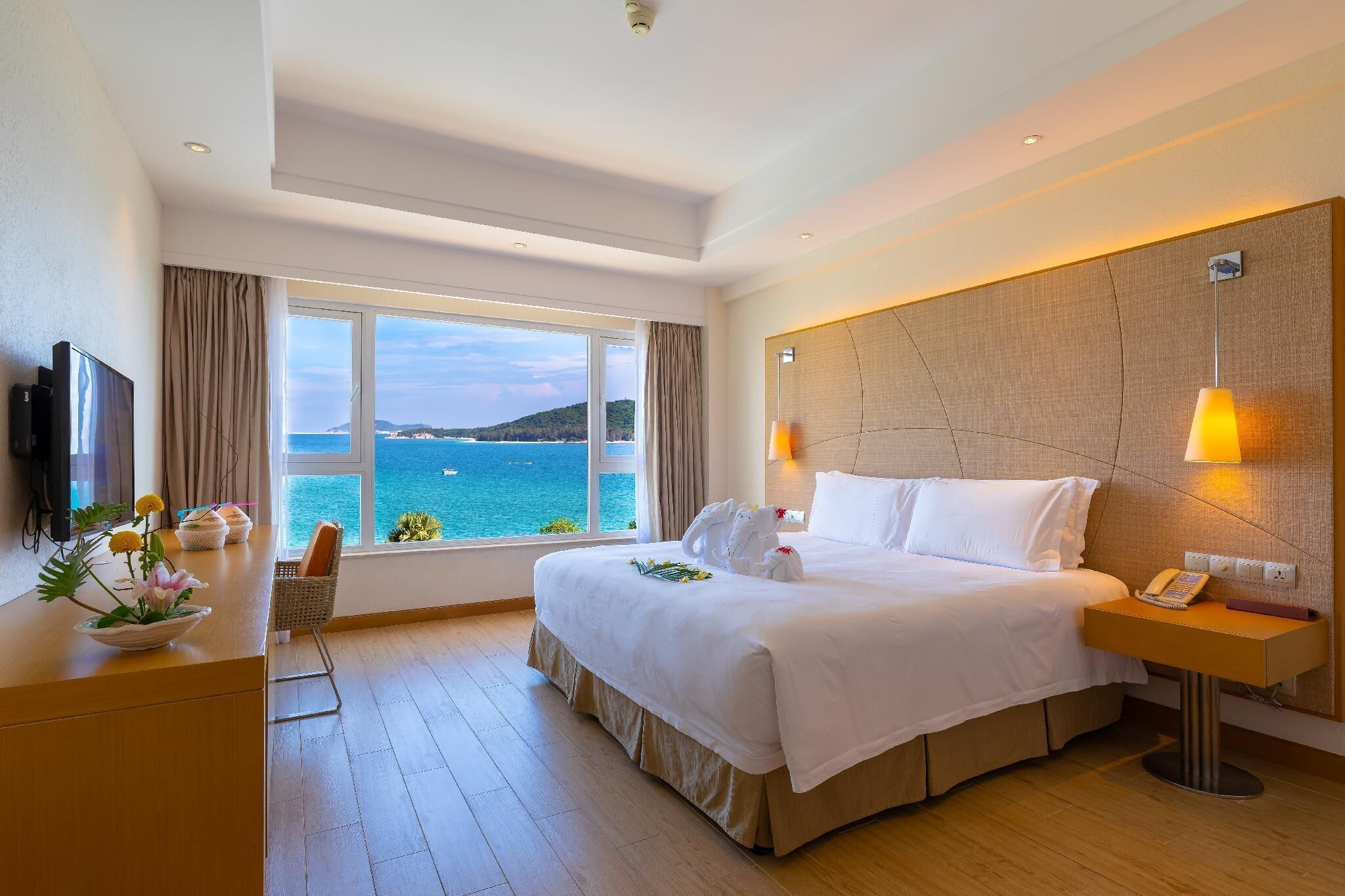 A hotel room with a large bed, a desk with a telephone, a TV, and a view of the sea.
