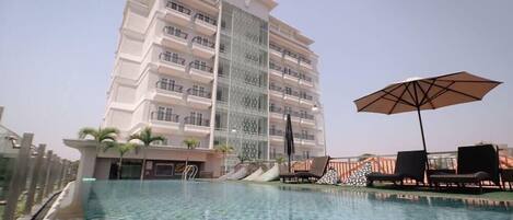 A modern hotel with a large pool, outdoor seating, and a tall building with balconies.