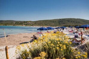 Une plage avec des transats et des parasols, une mer d’un bleu limpide et, en arrière-plan, un paysage vallonné.