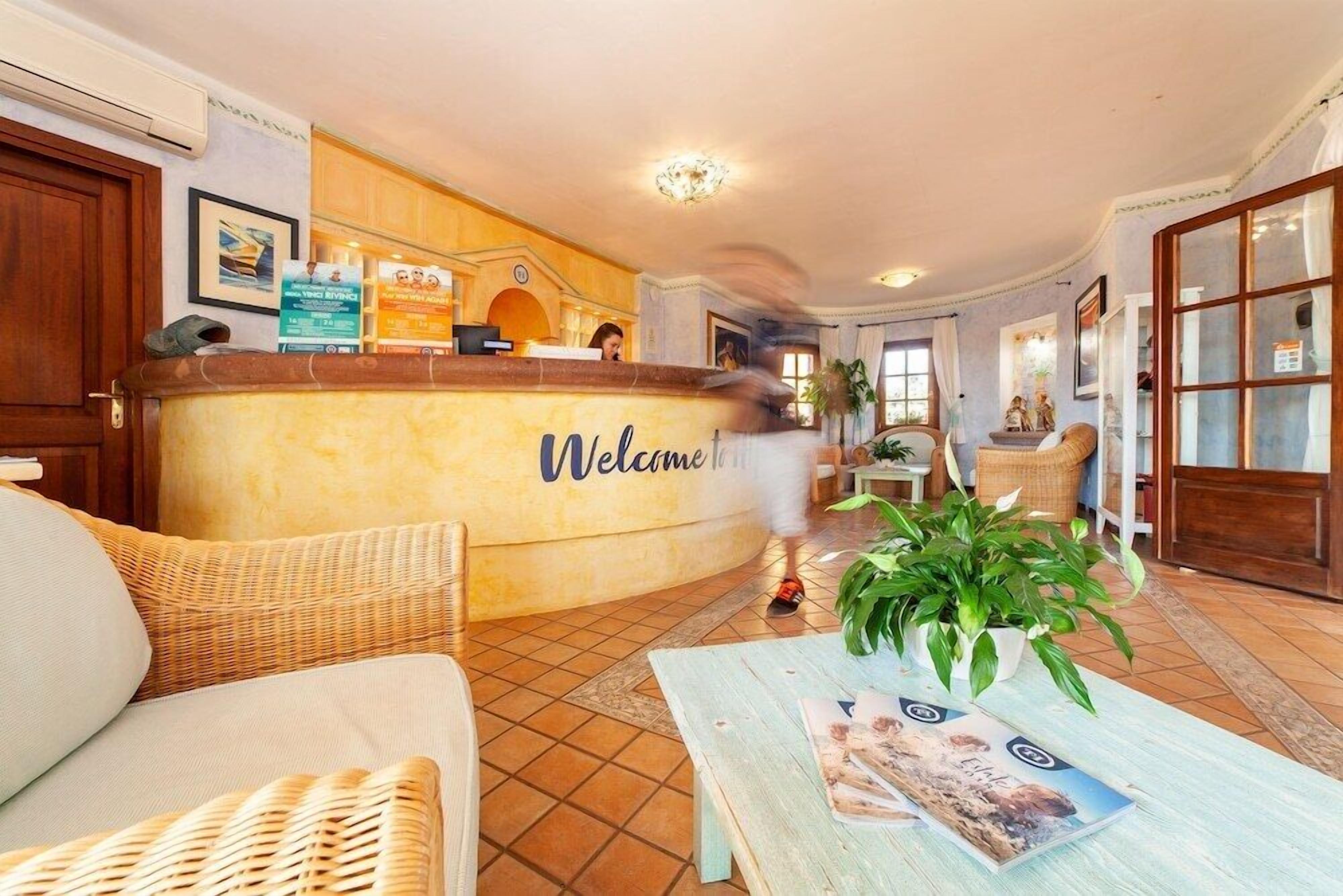 A hotel reception area with a 'Welcome' sign, wicker furniture, a tiled floor, and a reception desk.